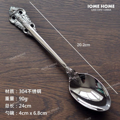 A silver-colored stainless steel spoon with an ornate vintage relief pattern on the handle.