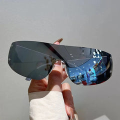 A hand holds a pair of rimless Y2K-style shield sunglasses with dark mirrored lenses and rivets.