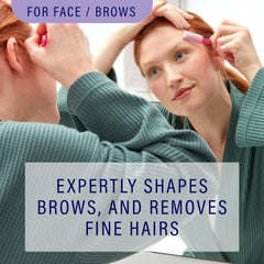 A woman uses a pink dermaplaning razor to shape her eyebrow while looking in a mirror.