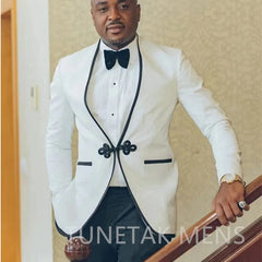 A man wears a beige two-piece tuxedo with black trim, a black bow tie, and a decorative clasp.