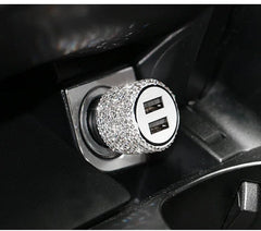 A dual USB car charger covered in sparkling rhinestones plugged into a vehicle power outlet.