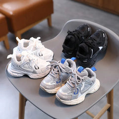 Three pairs of children's sneakers in white, black, and grey sit on a grey chair.