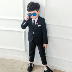 A young boy wears a black three-piece suit with a white shirt, black tie, and blue sunglasses.