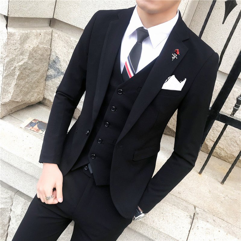 A man wears a slim-fit black three-piece suit with a white shirt, striped tie, and rose lapel pin.