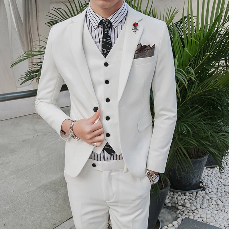 A man wears a white three-piece suit with a striped shirt, black tie, and a red lapel pin.