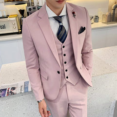 A man wears a slim-fit, light pink three-piece suit with a patterned tie and a rose lapel pin.