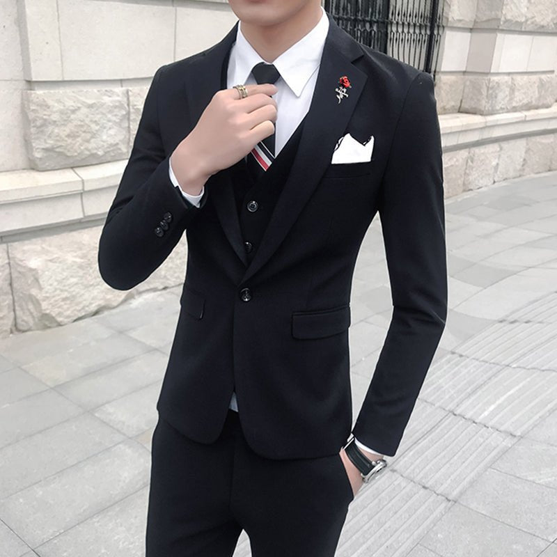 A man wears a slim-fit black three-piece suit with a white shirt, striped tie, and metal lapel pin.
