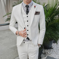 A man wears a white three-piece suit with a striped shirt, black tie, and a red lapel pin.