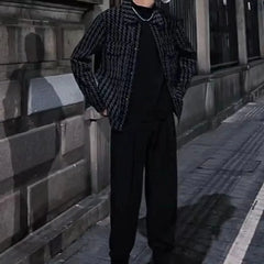 A man wears a black and white houndstooth patterned jacket with a square collar over a black shirt.