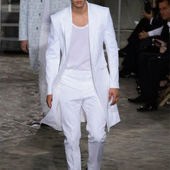 A man wears a white two-piece suit featuring a long blazer with a peaked lapel and matching pants.