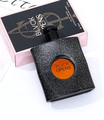 A black textured perfume bottle with an orange circular label sits in front of its pink box.