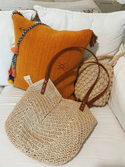 A straw shoulder bag with brown leather handles sits on a white surface next to a yellow pillow.