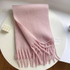 A soft, solid light pink winter scarf with long tassels, displayed on a white circular table.