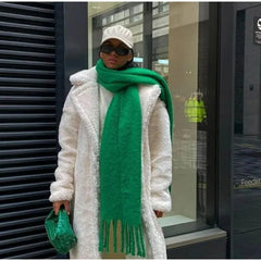A person wears a long, thick, solid green scarf with tassels over a white faux fur coat.