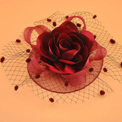 A burgundy rose fascinator hat with a polka dot mesh veil on a light orange background.