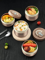Four round beige stainless steel lunch boxes are arranged on a dark surface, filled with various foods.
