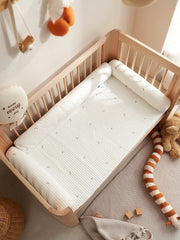 A white cylindrical crib bumper with a moon and star pattern inside a wooden baby crib.