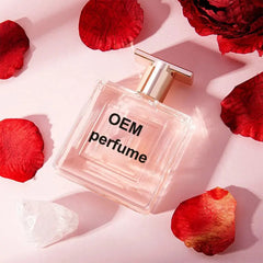 A square glass perfume bottle labeled OEM sits on a pink surface surrounded by red rose petals.