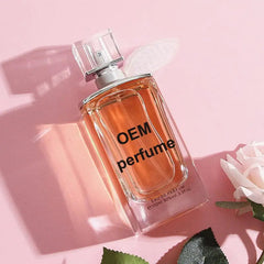 A rectangular glass perfume bottle with orange liquid sits on a pink surface with a rose.