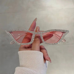 A hand holds cat-eye sunglasses with pink lenses and a frame encrusted with sparkling rhinestones.