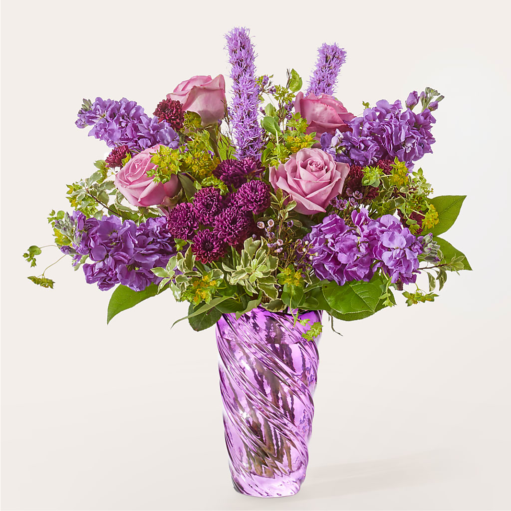 A bouquet of purple roses, stock, and liatris in a spiraled purple glass vase.