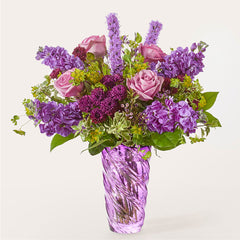 A bouquet of purple roses, stock, and liatris in a spiraled purple glass vase.