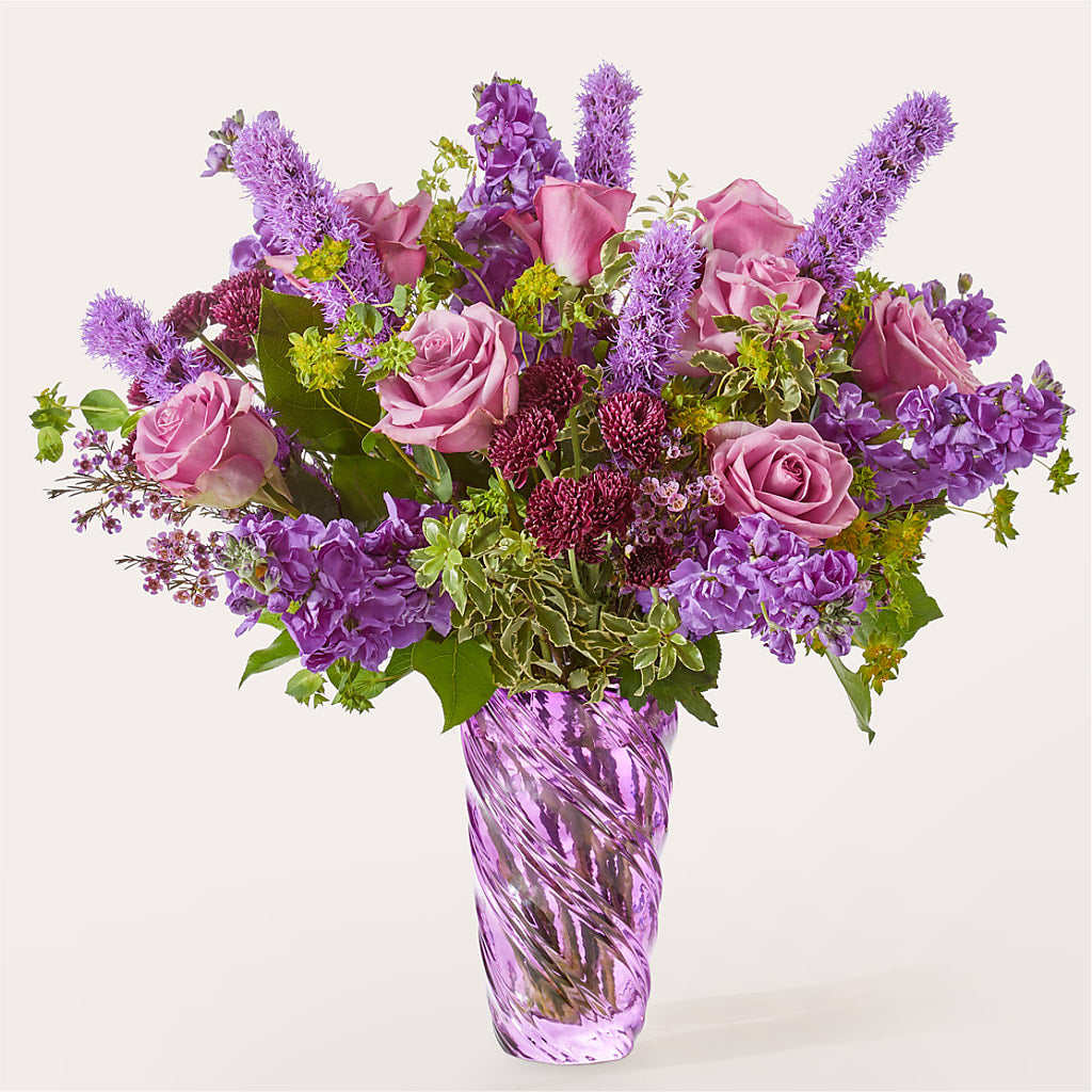 A bouquet of purple roses, stock, and liatris in a swirling purple glass vase.