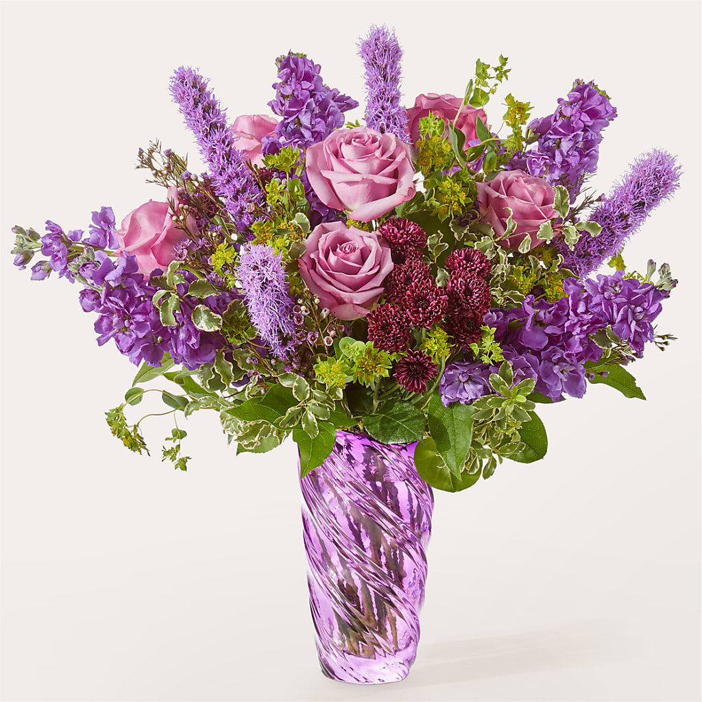A bouquet of purple roses, stock, and liatris in a textured purple glass vase.