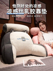 A grey and white plush bedside backrest cushion with a cute bear patch sits on a bed.