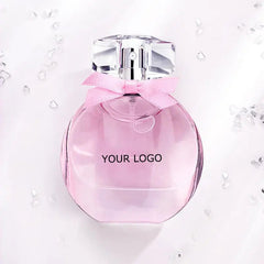 A round, pink Perfume Customize Luxury Fragrance bottle with a clear, faceted cap and light pink bow rests among small crystals on white, showcasing its delicate, feminine design. This long-lasting EDP is available for private label and OEM customization.