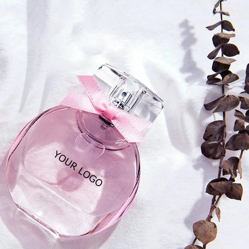A bottle of Perfume Customize Luxury Fragrance OEM Perfume Luxury Perfume Private Label Fashion Scent Long Lasting EDP with a pink design and clear cap sits next to dried leaves on a white surface.