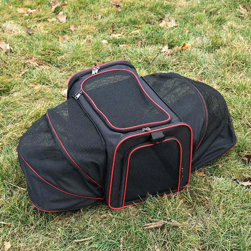 A black and red soft-sided pet carrier with expandable mesh sides sitting on grass.