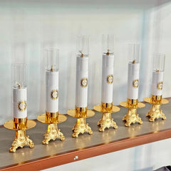 Six Catholic oil lamp candlesticks with gold bases and glass windshields on a shelf.