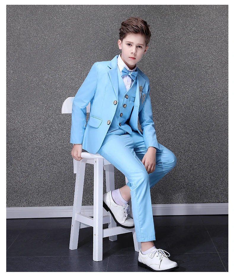 A boy wears a sky blue formal suit with a matching vest, bowtie, and white dress shoes.