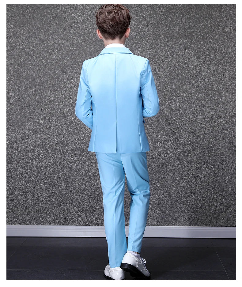 A boy stands facing away wearing a sky blue formal suit with a notched collar and white shoes.