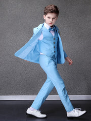 A young boy wears a sky blue formal suit with a matching vest, bowtie, and white dress shoes.