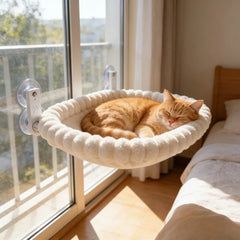 An orange cat sleeps in a round, plush, cream-colored hammock attached to a window by suction cups.
