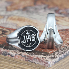A sterling silver ring featuring the JHS monogram and a cross on a textured black background.