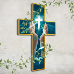 A wooden cross wall hanging featuring an illustration of hands raised toward a glowing cross.