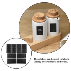 A set of black rectangular spice labels shown with two white ceramic jars on a wooden tray.