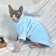 A Sphynx cat wears a light blue, long-sleeved fleece sweater with a high collar.