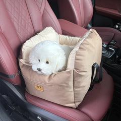 A small white dog sits inside a tan, cushioned pet car seat placed on a red leather car seat.