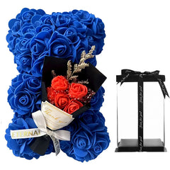 A blue artificial rose bear holding a small bouquet of orange flowers next to a clear gift box.