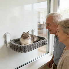 A grey plush cat hammock is mounted to a window with suction cups as an elderly couple watches.