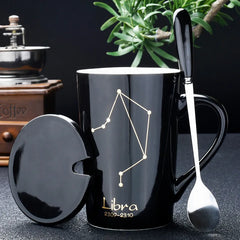 A black ceramic mug featuring a gold Libra constellation design, complete with a lid and spoon.