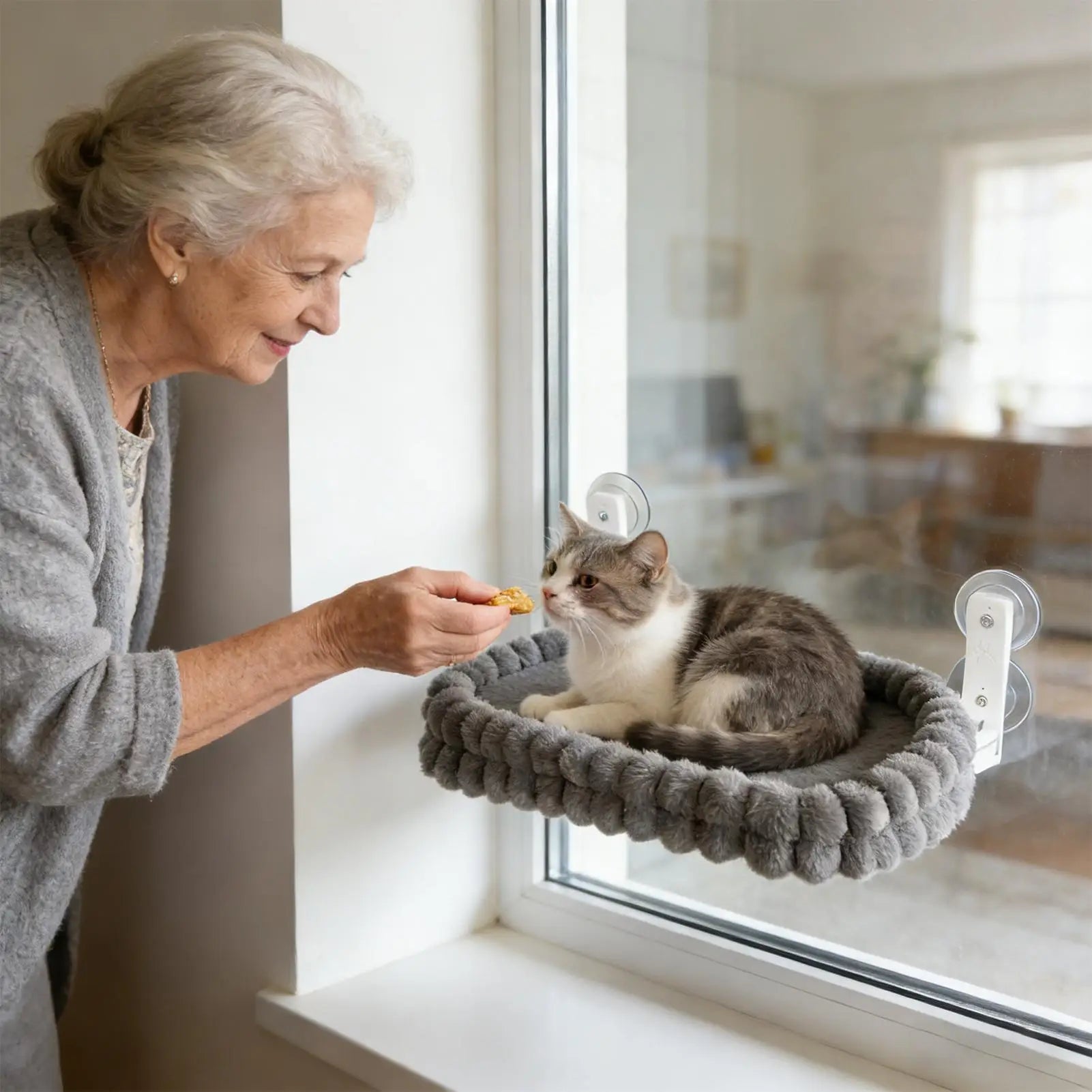 An elderly woman feeds a cat resting on a grey plush window perch attached with suction cups.