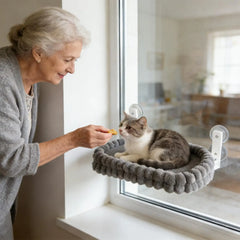 An elderly woman feeds a cat resting on a grey plush window perch attached with suction cups.
