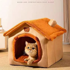 A tan and orange dog house style pet bed with a small chimney and a cat sitting inside.