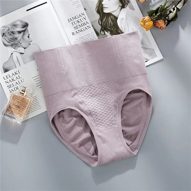 The Underwear Bragas Fajas Women Shaper Thong Bodyshaper Nude Low High Waist Pant Tummy Tuck Shapewear Panty in light mauve with a textured panel is displayed on magazines beside perfume and orange flowers on a gray surface.