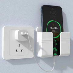 A white wall-mounted mobile phone holder holding a charging smartphone next to a wall outlet.